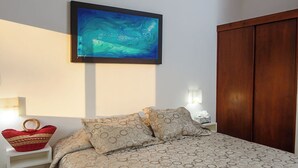 Une chambre à coucher bien rangée, avec un lit, un climatiseur fixé au mur, un ventilateur de plafond, une œuvre d’art encadrée, une lampe de chevet et une armoire en bois.