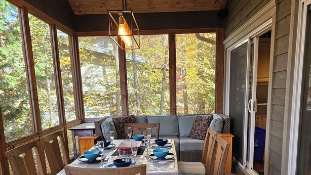 A covered outdoor dining area with a wooden table and chairs, a blue and white patterned rug, and a view of trees through large windows. - Lakefront Spectacle In Beaver Valley / Lake Eugenia (Flesherton)