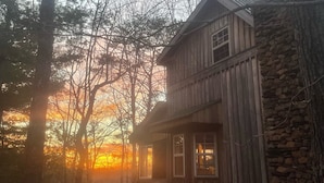 A rustic wooden cabin with a stone wall, surrounded by trees, during sunset.
