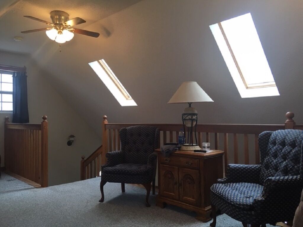 Ein Zimmer mit schrägen Wänden, Dachfenstern, zwei gepolsterten Sesseln, einem Holztischchen mit Lampe und einer Treppe.