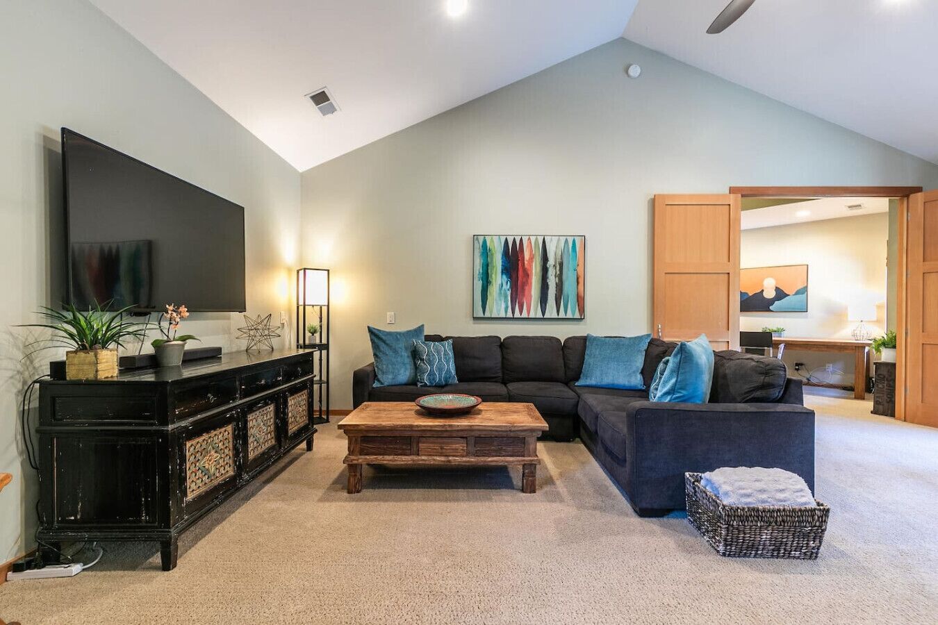 Un salon comprenant un meuble de rangement noir pour appareils électroniques, une table basse en bois, un canapé foncé agrémenté de coussins bleus et une peinture abstraite aux couleurs vives accrochée au mur.