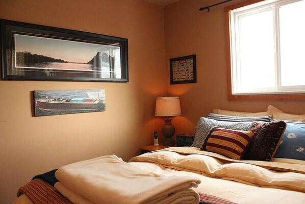 Un dormitorio con cama, almohadas, mesita de noche con lámpara y cuadros en la pared.