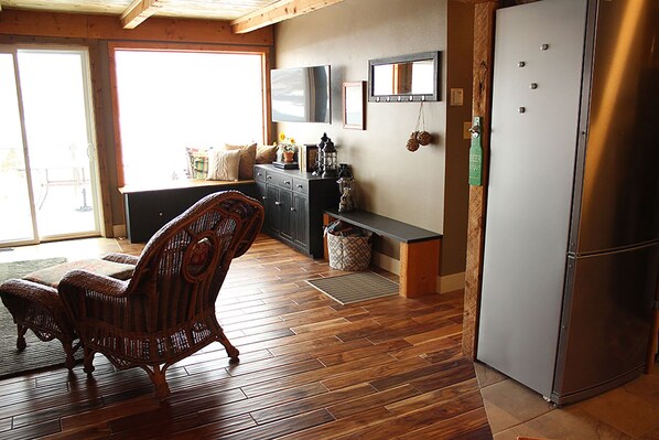 Una cocina con piso de madera, una silla de mimbre, un refrigerador y un banco con diversos objetos encima.