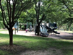 Children can play on the slides, climbing structures, and swings in this shaded playground area surrounded by trees and grass.