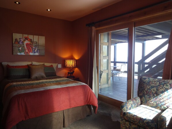 A bedroom with a large bed, a chair, a painting, and a window with a view of an outdoor deck.