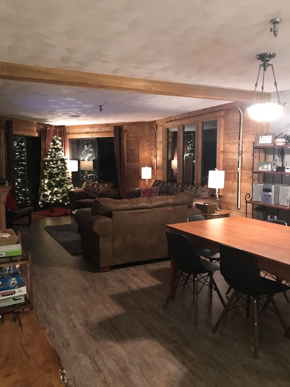A cozy living room with a wooden ceiling, a Christmas tree, a sofa, a dining table with chairs, and a kitchen area with appliances.