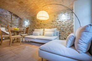 A cozy living room with a stone wall, a white sofa, a wooden dining table with chairs, and a staircase.
