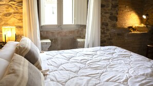 Une chambre avec un mur en pierre, un lit avec une literie blanche, une table de chevet avec une lampe et une fenêtre avec des rideaux.