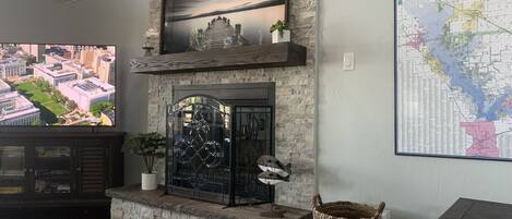 A living room with a stone fireplace, a mounted television displaying a cityscape, and a map on the wall.
