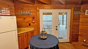 A wooden cabin interior with a kitchen area, a dining table with a tray, a refrigerator, and a door leading to an outdoor deck. - Log Cabin with large deck and dock in best location on the Lake! (mccormick)