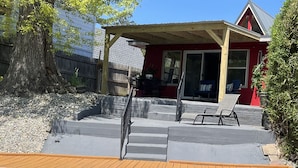 Um deck de madeira com área de pátio de concreto, espaço coberto para sentar e um edifício vermelho ao fundo.