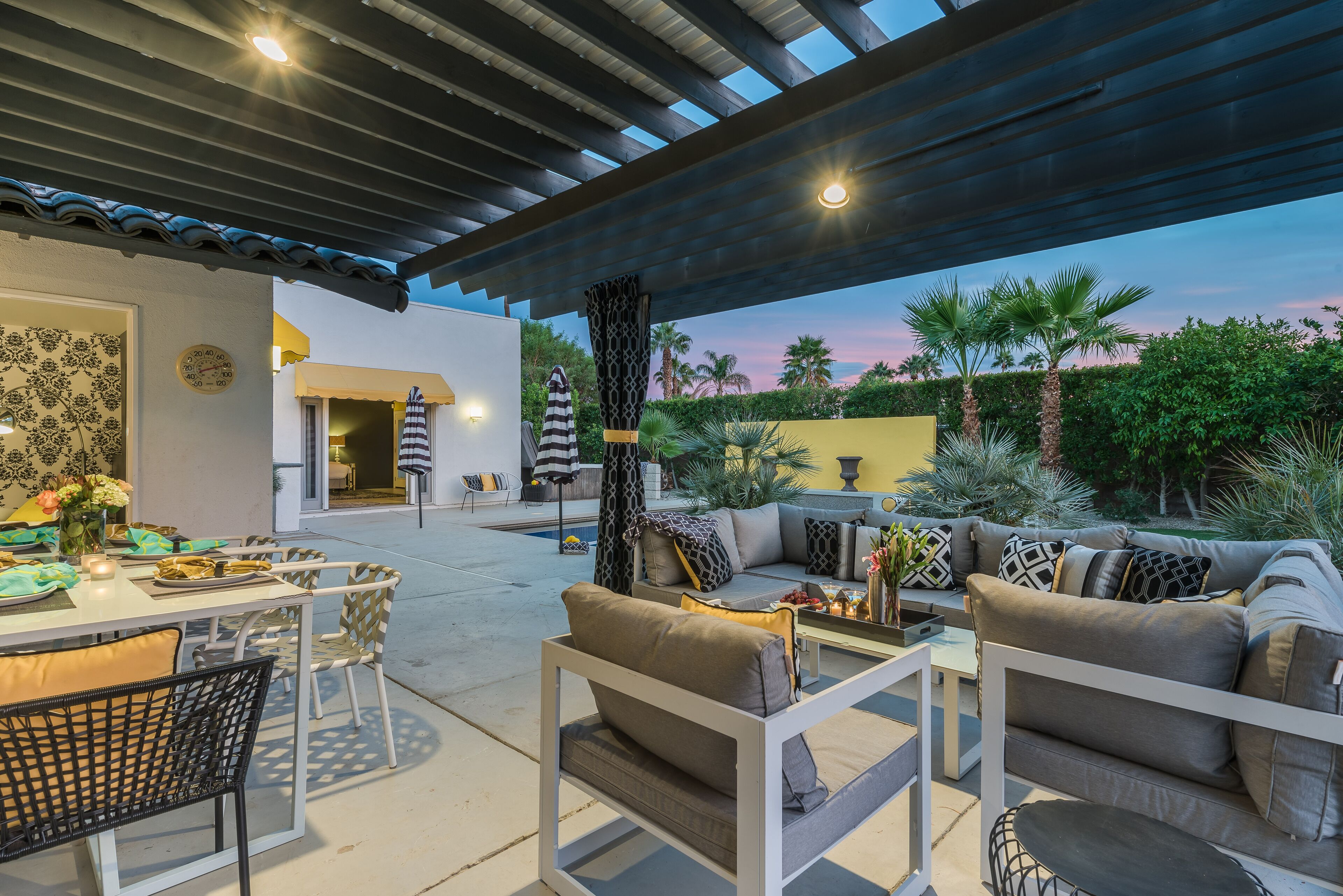 An outdoor patio area with a covered seating area, a dining table, and a pool in the background.