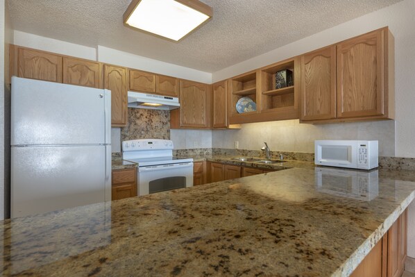 A kitchen with granite countertops, wooden cabinets, a refrigerator, stove, microwave, and a dish rack.