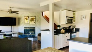 Uma sala de estar moderna com televisão de tela plana, lareira, área de jantar com mesa de madeira e cozinha com armários brancos e micro-ondas.