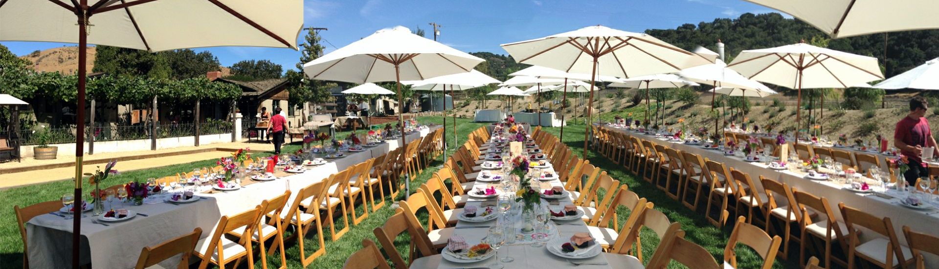 Un espace de restauration en plein air avec plusieurs tables recouvertes de nappes blanches et entourées de chaises en bois, le tout protégé par de grands parasols.