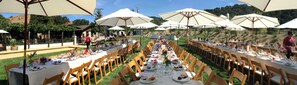 An outdoor dining area with multiple tables set with white tablecloths and wooden chairs, surrounded by large umbrellas.