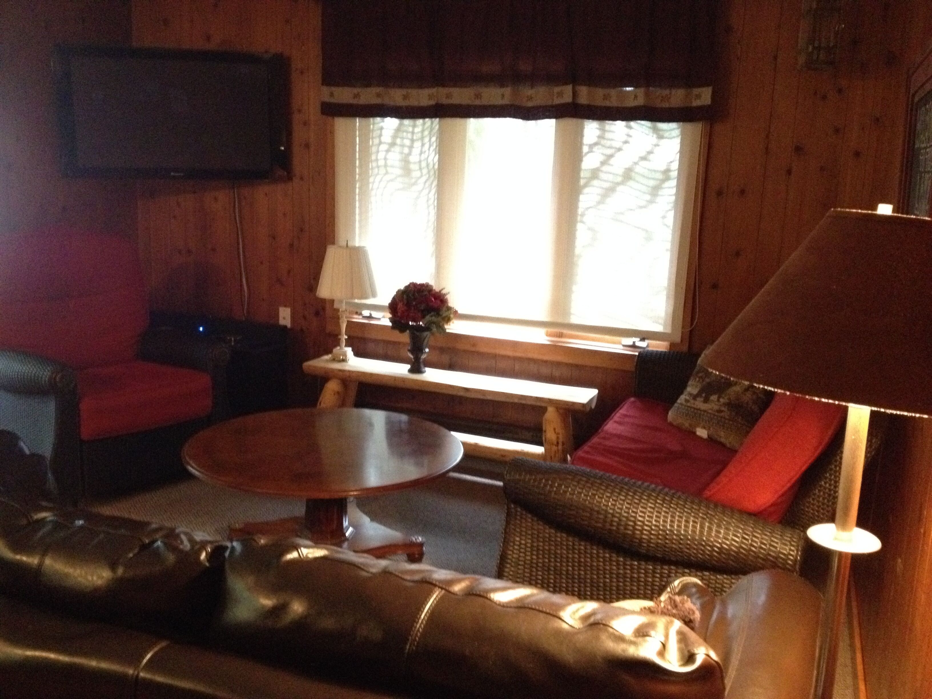 A cozy living room with a leather sofa, a wooden coffee table, a red armchair, and a bench. A lamp and a vase with flowers are on a small table.