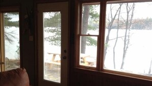 Ein Interieur einer Holzhütte mit Holzboden, braunem Sofa, einer Holzbank und einem großen Fenster mit Blick auf eine verschneite Landschaft.
