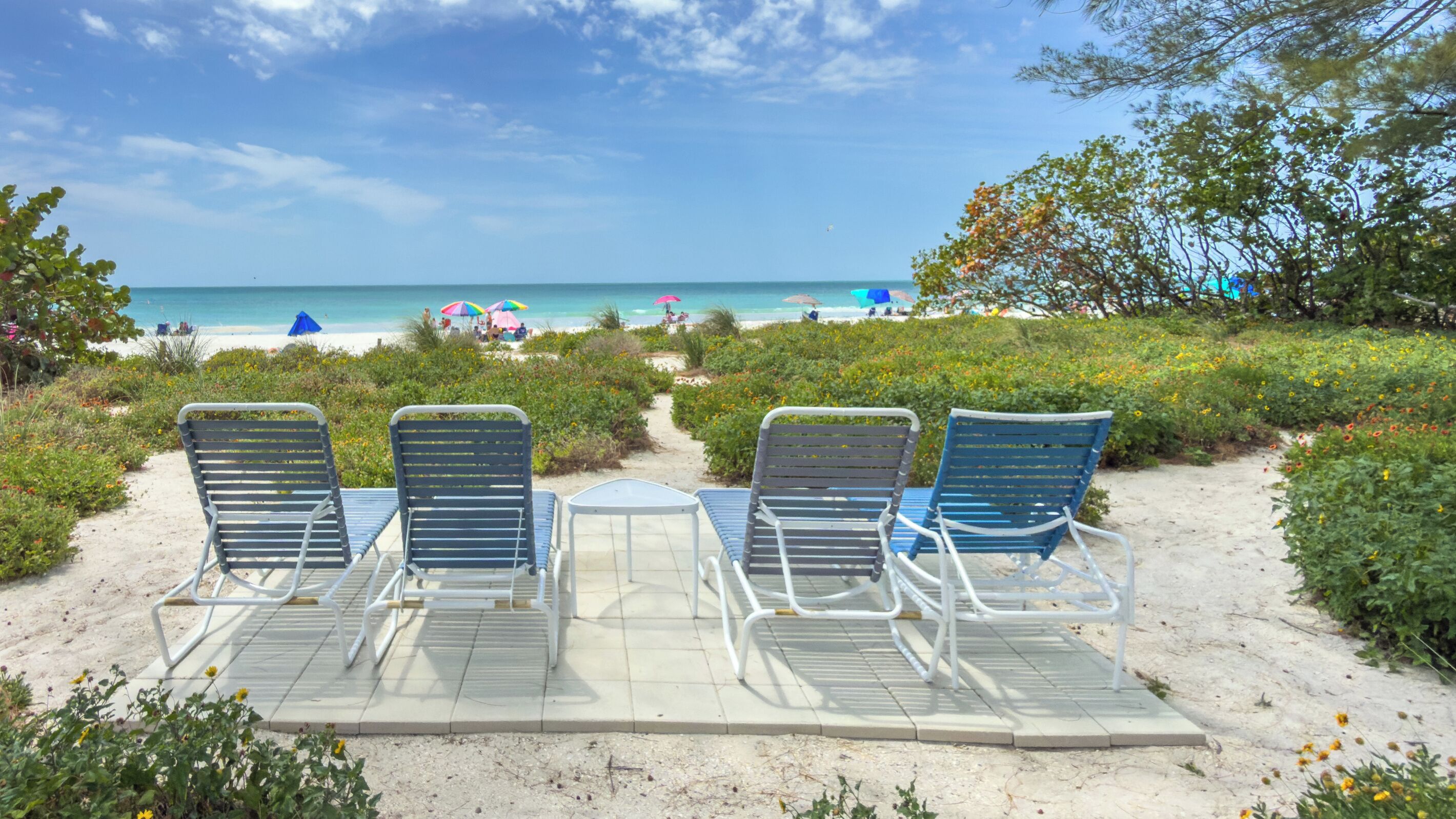 Un espace aménagé avec des chaises et une table en métal, situé au bord de la plage et offrant une vue sur une plage de sable, des parasols et des personnes au loin.