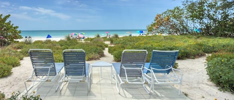 A beachside seating area with metal chairs and a table, overlooking a sandy beach with umbrellas and people in the distance.