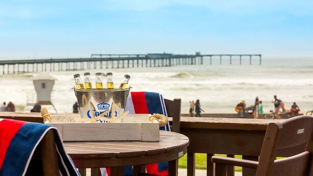 Ein Tisch am Strand mit einem Eimer voller Bierflaschen, einem Holenstuhl und Blick auf das Meer und den Pier.