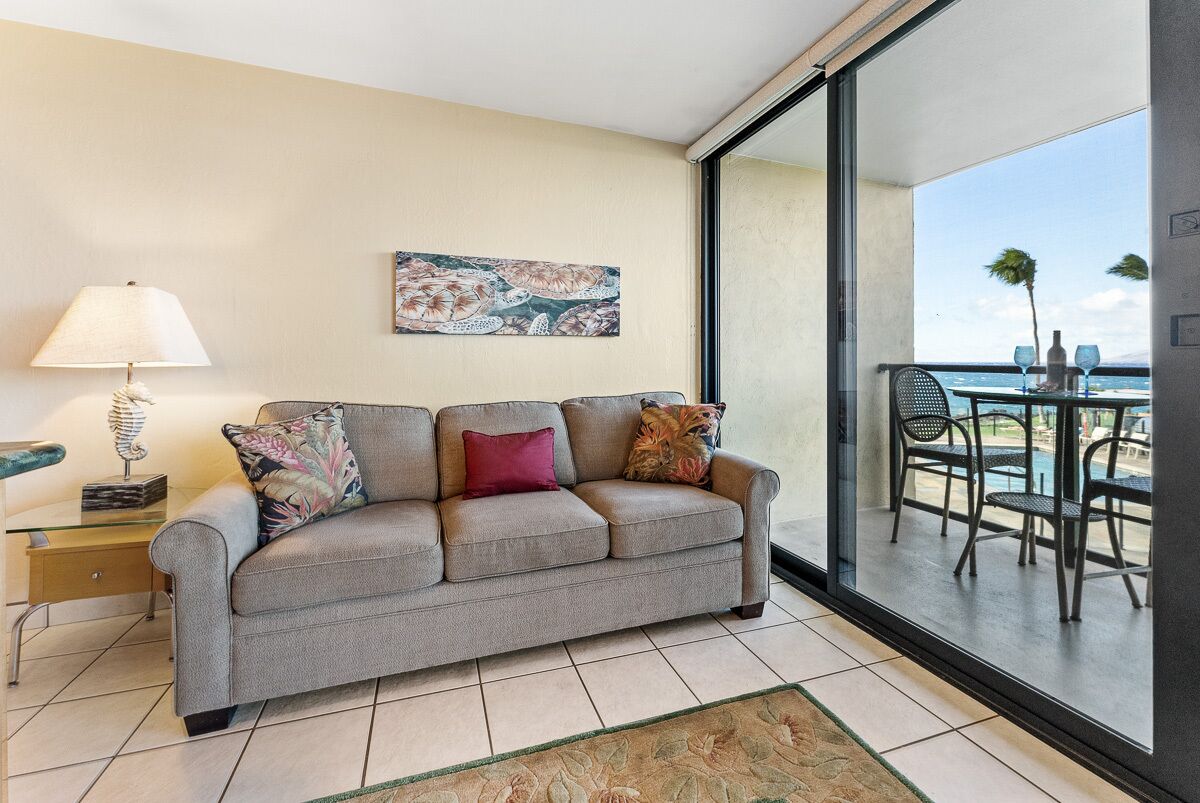 A modern living room with a grey sofa, a glass coffee table, and a floor lamp. A large window offers a view of a balcony with outdoor furniture.