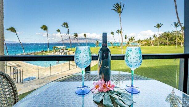 Mesa con dos copas de vino y una botella, con vistas a una piscina y una playa con palmeras y cielo despejado.