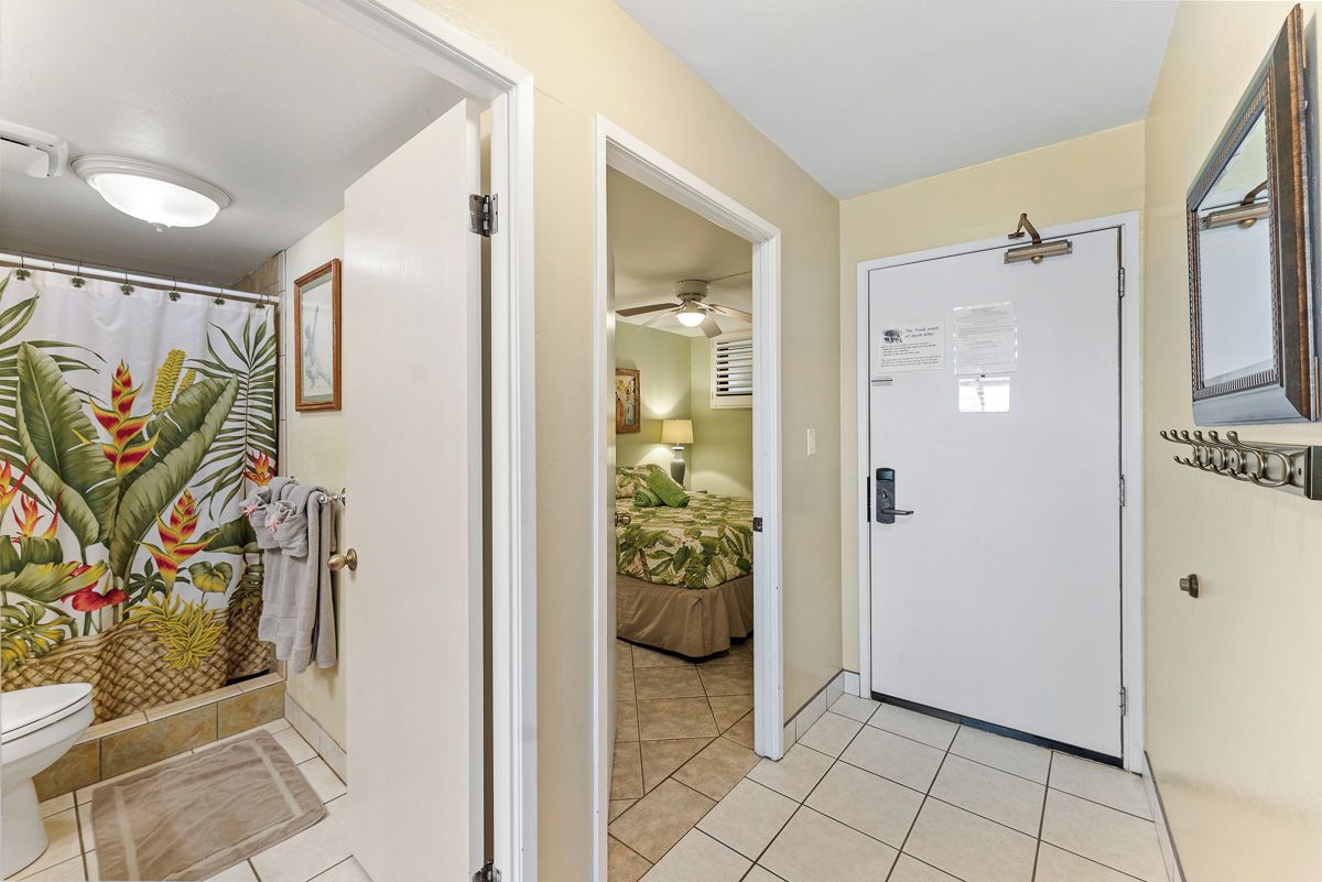 A hotel hallway with a bathroom featuring a tropical mural, a bed in the adjacent room, and a door with a bulletin board.
