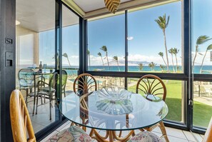 A glass-enclosed dining area with a table and chairs, overlooking a beach with palm trees and a clear sky. - 207 - Lovely Oceanfront One Bedroom Unit! (Kihei)