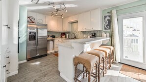 A modern kitchen with a stainless steel refrigerator, a central island with bar stools, and a view through a large window. - Almost Beachfront- Beautiful Pirates Beach home- Sleeps 14-Steps to beach!! (Galveston)