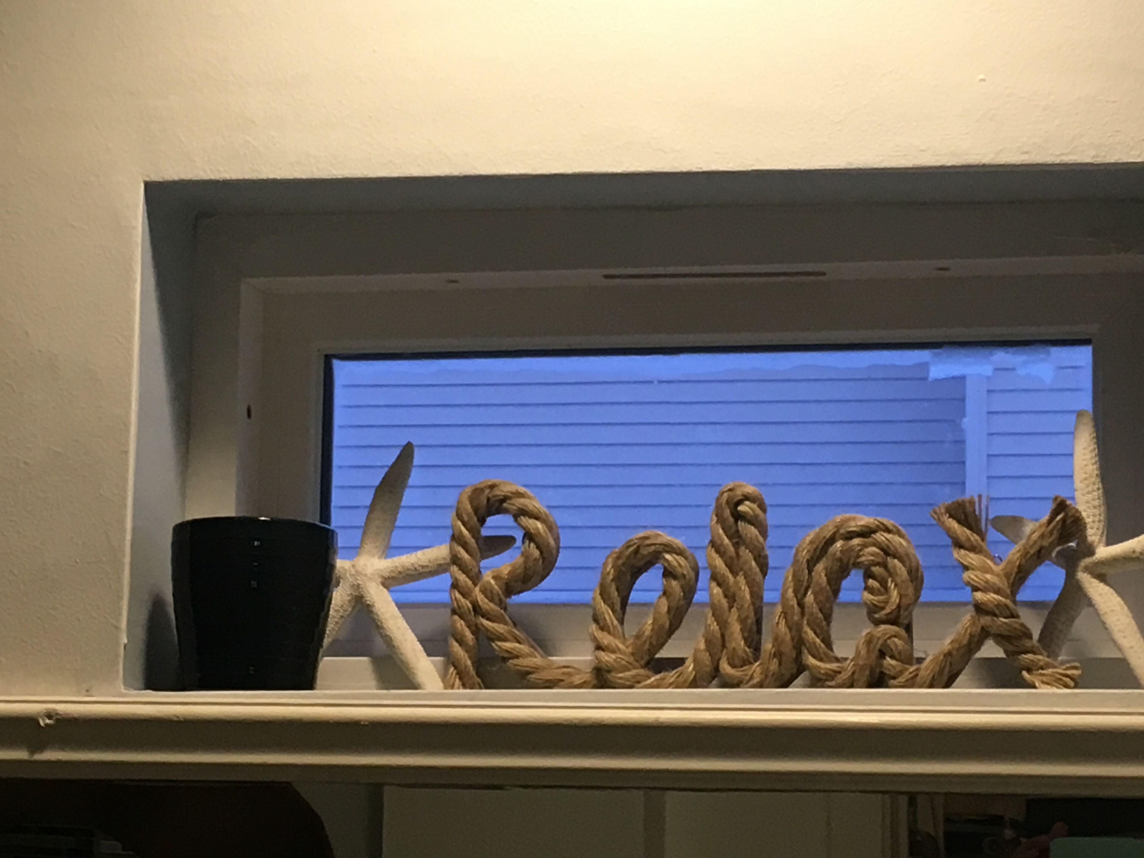 Une fenêtre ornée d’une décoration faite de cordes formant le mot « relax », et un objet noir posé sur le rebord.