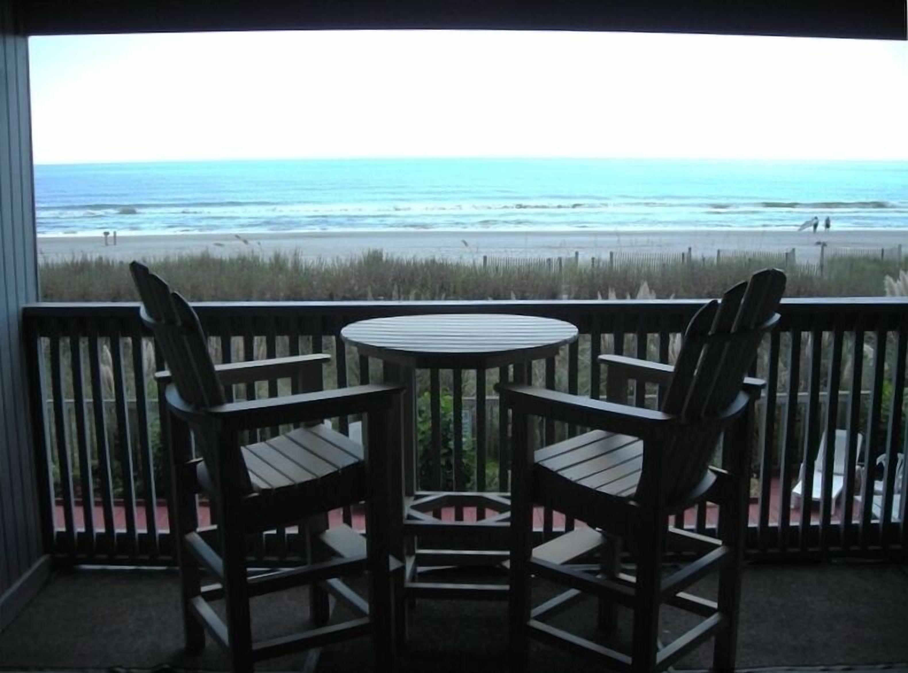Een balkon met twee houten stoelen en een klein, rond tafeltje, met uitzicht op een strand, de oceaan en een verre kustlijn.