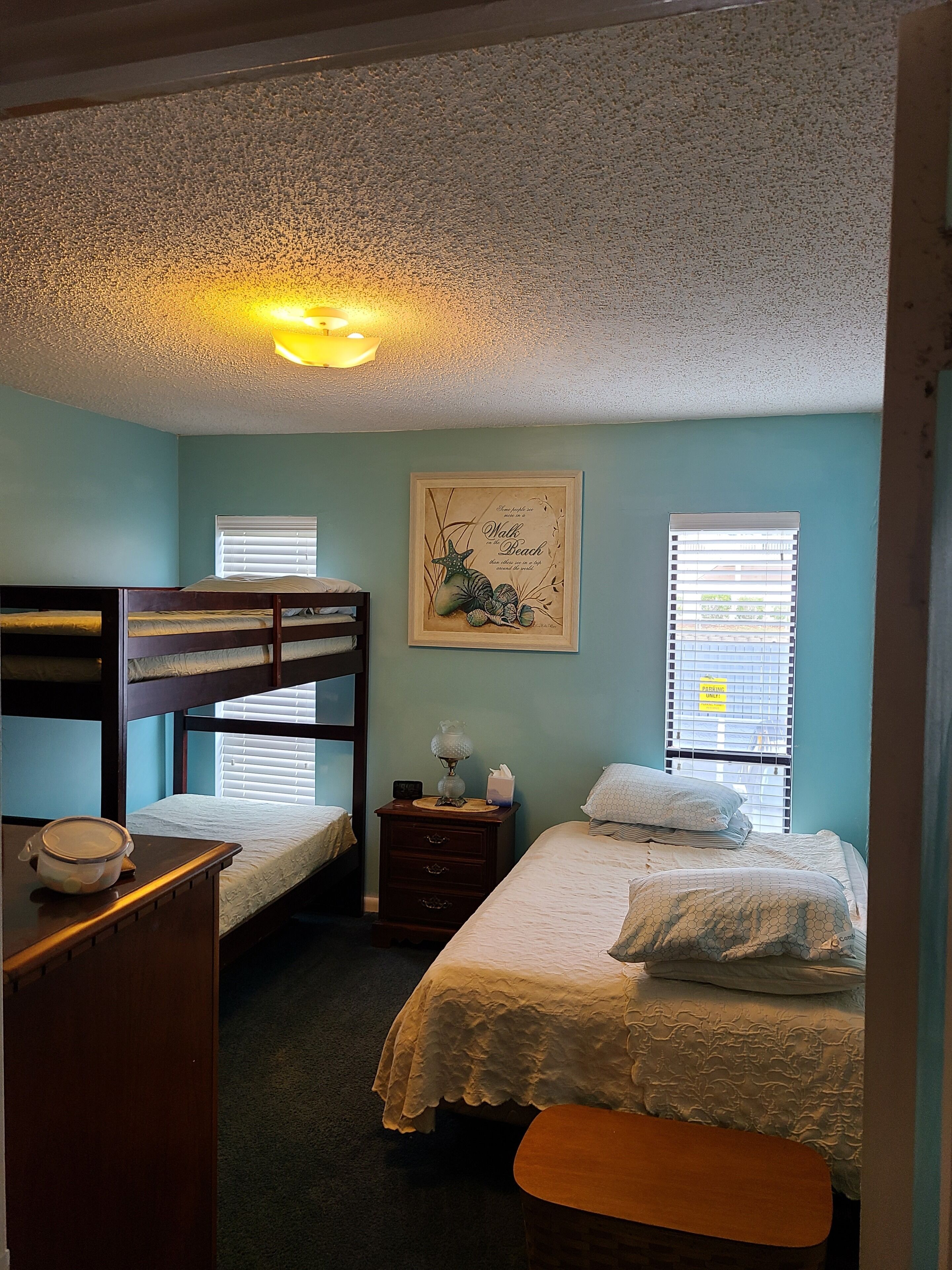 A room with bunk beds, a bedside table with a lamp, a framed picture on the wall, and a window with blinds.