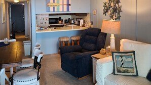 Ein Wohnzimmer mit einem dunkelblauen Sofa, einem weißen Sessel und einem weißen Couche. Es gibt eine Küchenzeile mit Mikrowelle, Herd und Spüle. Über der Küche ist ein Deckenventilator zu sehen.