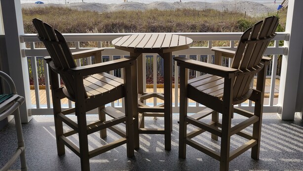 Ein Balkon mit zwei Holzstühlen und einem kleinen Tisch, mit Blick auf einen Strand und Wellen, sowie Menschen in der Ferne.