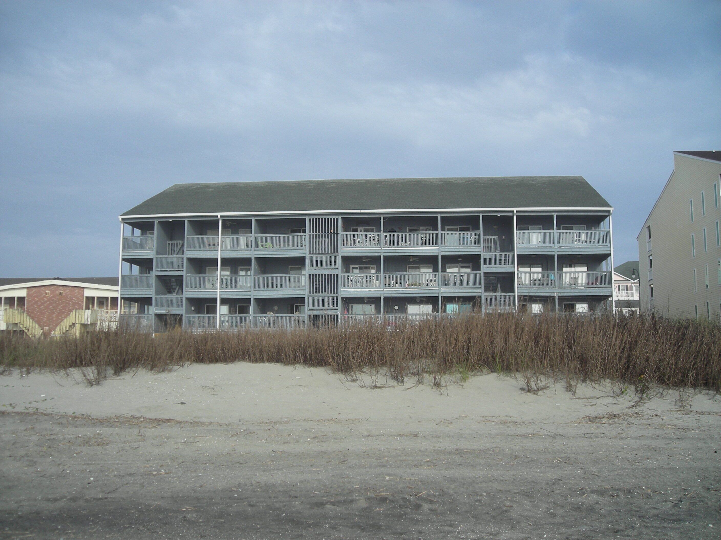A multi-story building with balconies, located near a sandy area with vegetation.