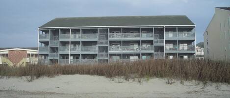 A multi-story building with balconies, located near a sandy area with vegetation.