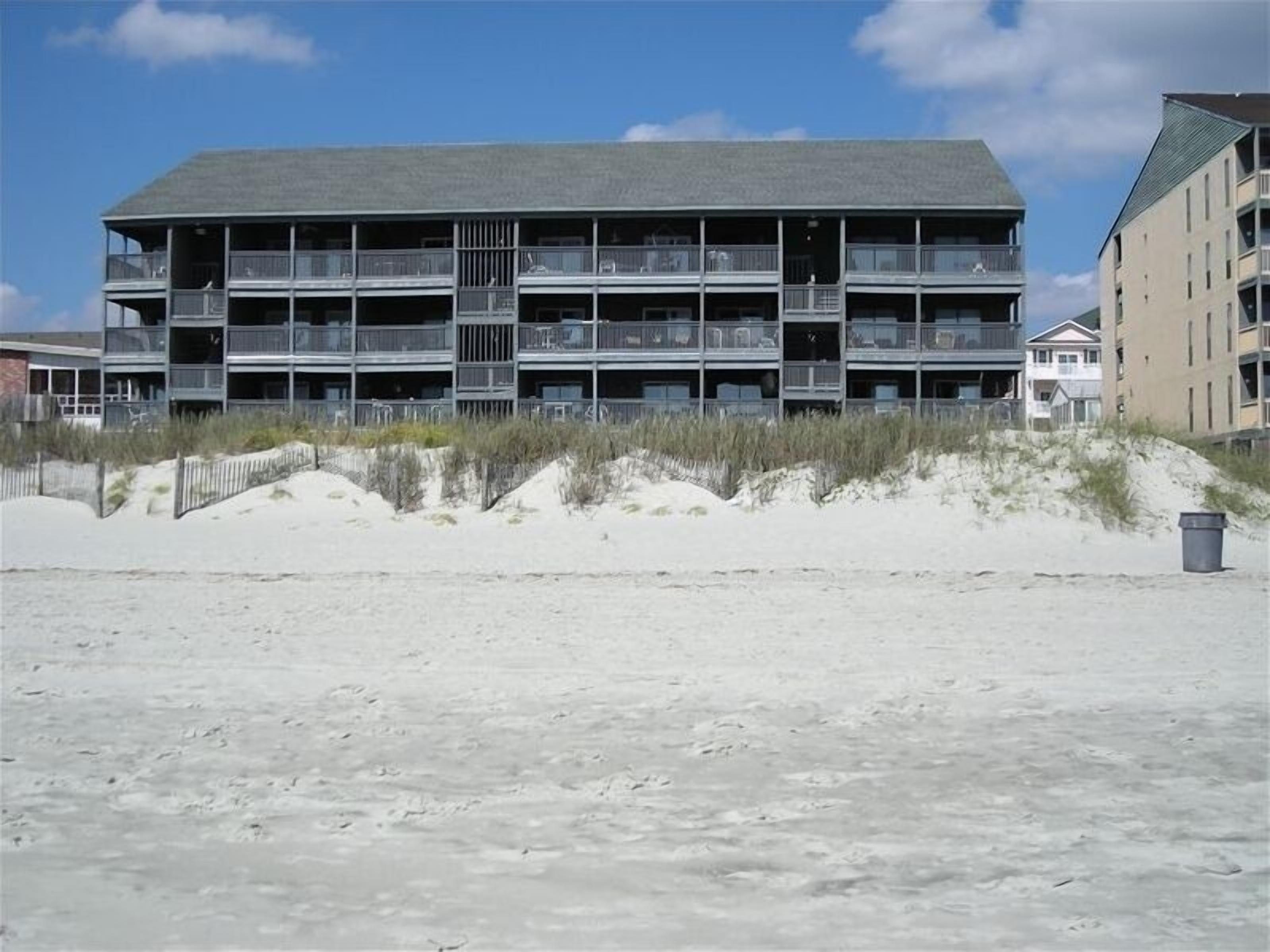 A multi-story hotel with balconies, located on a sandy beach with dunes in the background.