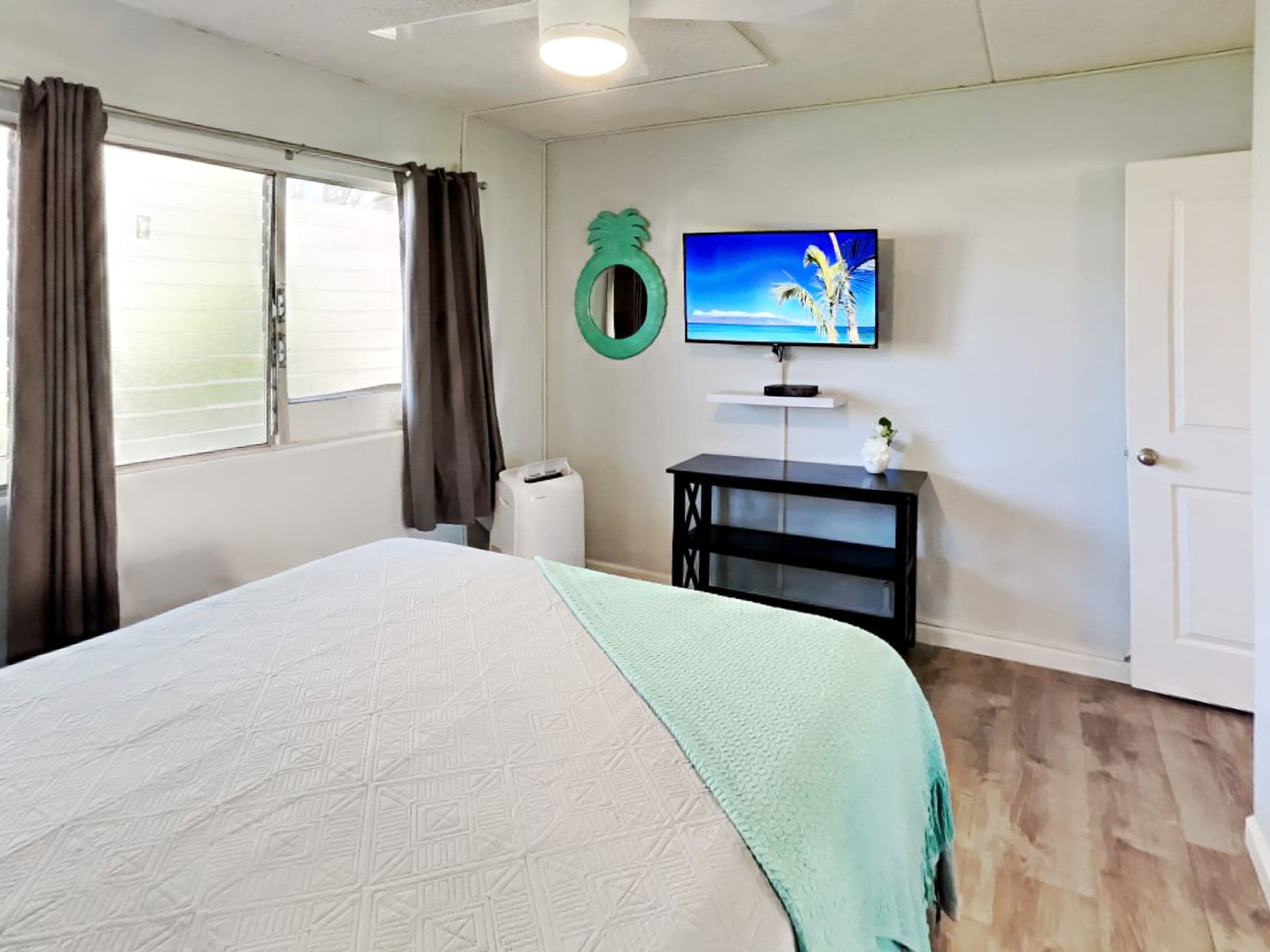 A bedroom with a bed, a TV, a small black table, a window with curtains, and a door.