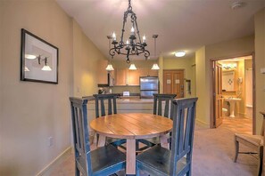A dining area with a wooden table and chairs, a chandelier, and a kitchen in the background.