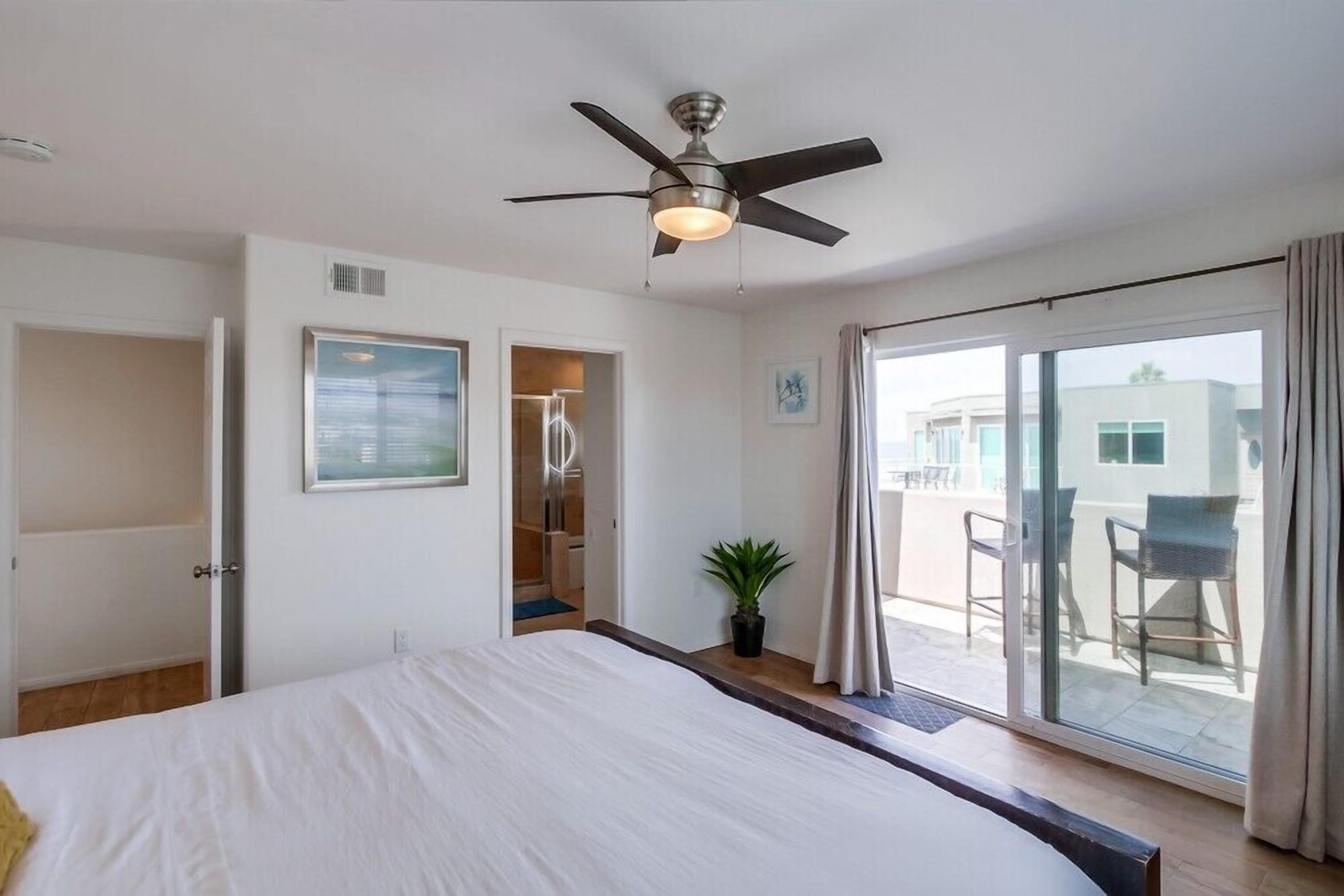 A hotel room with a bed, ceiling fan, sliding glass door to a balcony, and a potted plant.