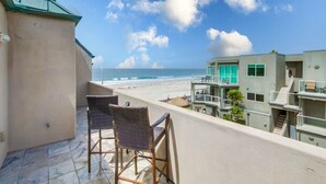 Uma varanda com duas cadeiras de vime, com vista para uma praia e um edifício com janela verde.