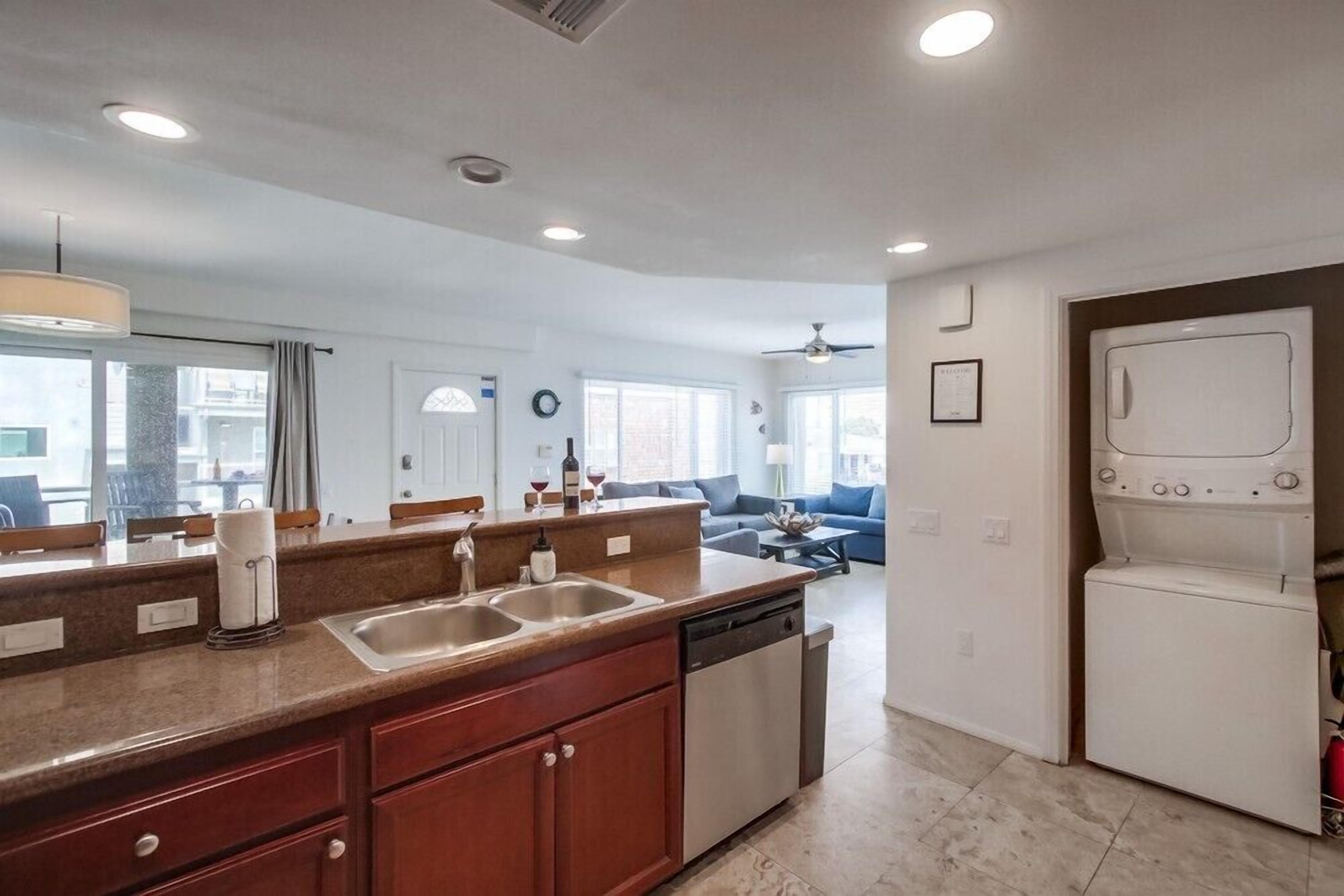 A modern kitchen with a double sink, dishwasher, and a built-in oven. There is a dining area with a sofa and a coffee table in the background.