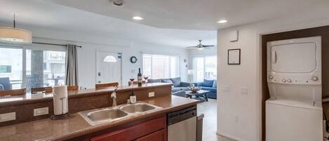 A modern kitchen with a double sink, dishwasher, and a built-in oven. There is a dining area with a sofa and a coffee table in the background.
