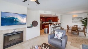 Uma sala de estar moderna com lareira, televisão exibindo uma cena de surf, ventilador de teto e área de jantar com mobiliário de madeira.