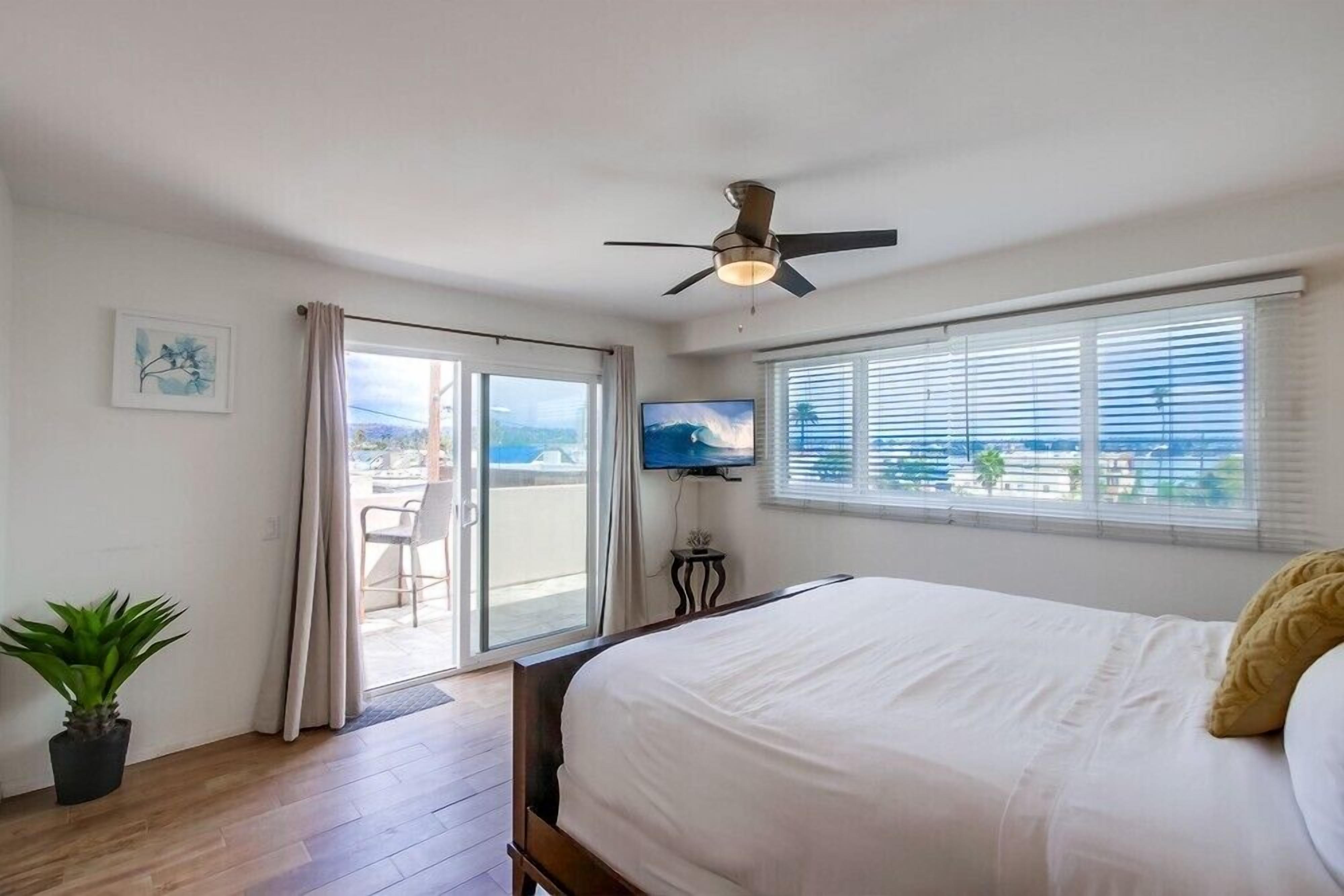 A bedroom with a bed, a ceiling fan, a TV, and a balcony with a view.