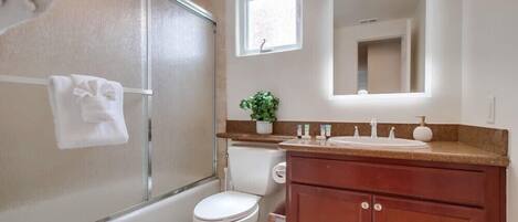 Een badkamer met een douche in een glazen cabine, een wit toilet, een wastafel met spiegel en een klein raam.