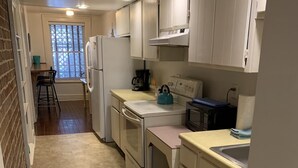 A narrow kitchen with white cabinets, a refrigerator, a stove, and a sink. There is a window with blinds and a chair in the background. - Budget Friendly Apartment on Jones Street! (Savannah)