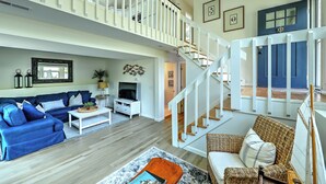 A spacious living area with a blue sofa set, a white coffee table, and a wicker chair. A staircase with white railings leads to an upper level. - Fantastic Contemporary ,Min to Narr beach. (Narragansett)