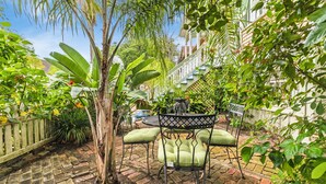 A patio with a round table and chairs, surrounded by lush greenery and a brick floor.